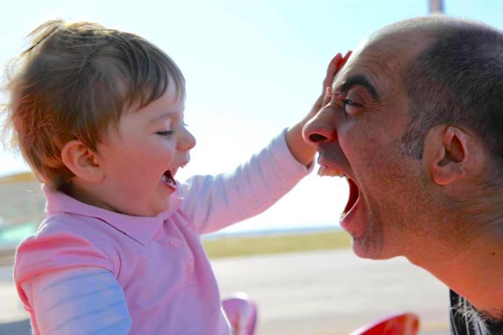 Pai e filha a brincar ao ar livre: ele de boca aberta, ela a rir com a mão na cara dele
