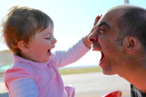 Pai e filha a brincar ao ar livre: ele de boca aberta, ela a rir com a mão na cara dele
